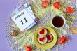 Calendar for February 11: the name of the month February in English, the numbers 11 on the loose-leaf calendar, a cup of tea, heart-shaped cookies, physalis branches on a yellow openwork napkin