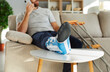 Close up of injured man with bandage on leg sit on sofa talk on cellphone with doctor. Unhealthy guy with sling on broken foot suffer from fracture or injury. Rehabilitation at home concept.