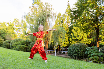 Cute little caucasian girl seven years old in red sport wushu uniform exercising in park at summer day. Lifestyle portrait of kung fu fighter child athlete