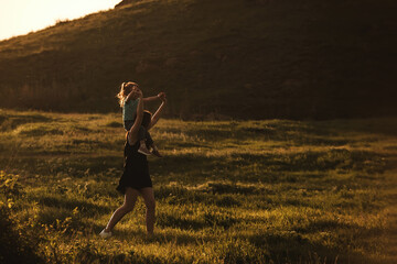 Two sisters of different ages have fun and actively spend time on a walk, the youngest sits on the shoulders of the older girl and they run on the grass in the rays of the setting sun