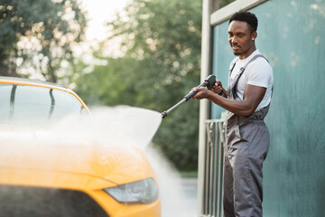 Wall Mural - Handsome African young man in protective overalls, washing his yellow car manually with water high-pressure hose at outdoor self wash service. Car washing concept.