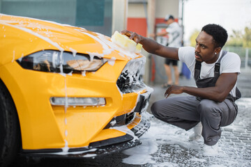 Poster - Car detailing wash at outdoors car wash service. Handsome African American man in white t-shirt and overalls washing car radiator grille and hood of his new yellow sport car with foam and sponge