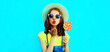 Summer portrait of beautiful young woman with lollipop blowing her lips sending sweet air kiss wearing a straw hat on blue background