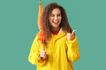 Poster - Young African-American woman in yellow raincoat with umbrella showing thumb-up on green background