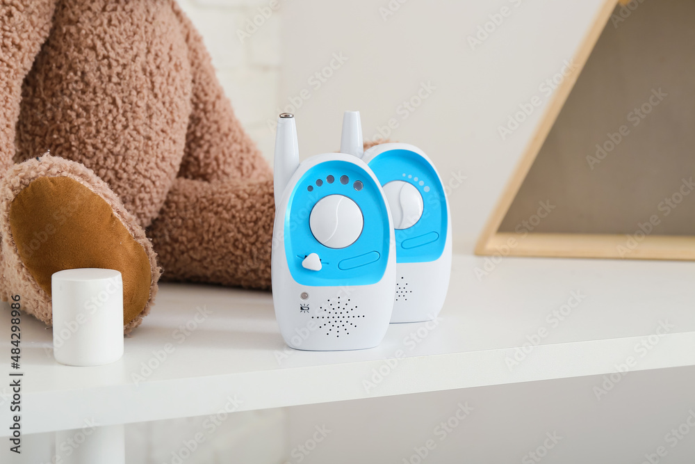 Modern baby monitors and toy bear on shelf near light wall