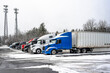 Big rigs semi trucks with different semi trailers standing for truck driver rest on the winter truck stop parking lot with snow and ice surface