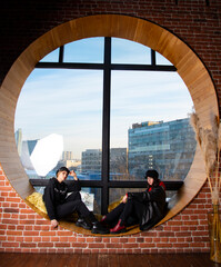 Two teenage girls are sitting on a wide windowsill by a large round window.