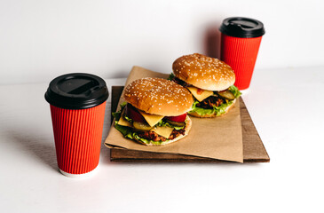 Two large burgers on kraft paper and a wooden board, with two red paper cups of coffee on a white background, a wooden table. Fast junk American food. Large portions. Takeaway delivery. Vegetarian.