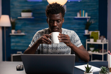 Poster - Businessman holding cup drinking coffee while reading business project using laptop computer working remote from home. Freelancer man sitting at desk during online lesson in living room