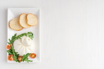 Wall Mural - Top view of homemade organic burrata Italian milk cheese  made of mozzarella and cream served in rectangular plate with cherry tomatoes, bread and arugula on white wooden background with copy space