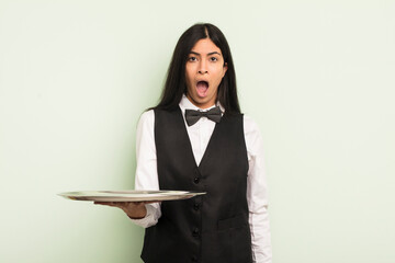 Wall Mural - young pretty hispanic woman looking very shocked or surprised. waiter with tray concept