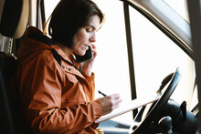 Woman Talking On Smartphone And Writing Notes On Clipboard In Truck