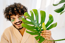 Portrait Of Attractive Young Asian Man With Green Eye Patches In Bathrobe Holding Big Green Monstera Leaf On White Background. Cosmetology Beauty, Ecology Lifestyle Concept.