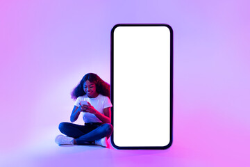 Wall Mural - Cheery black woman sitting near giant smartphone with mockup for your app design, using mobile device in neon light