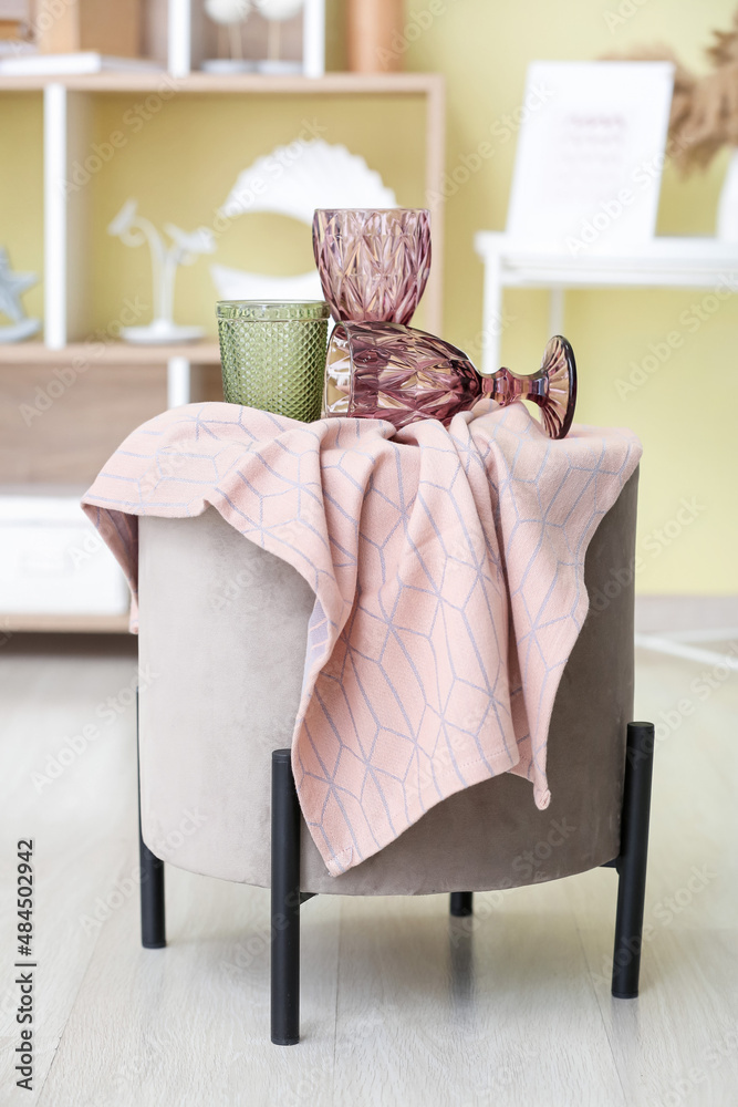 Different stylish glasses and napkin on pouf in room