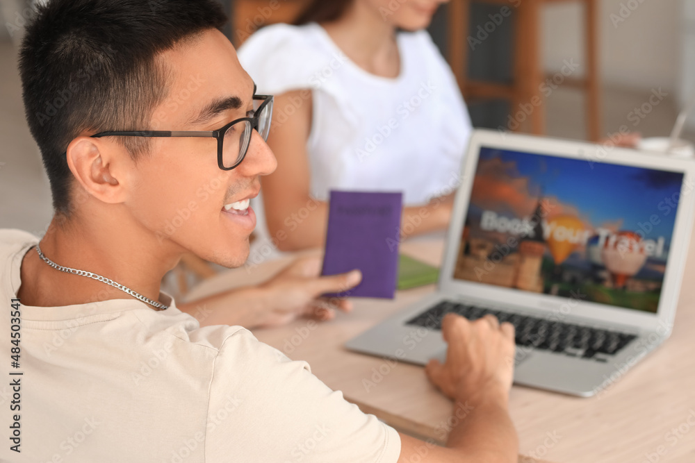 Young man with passport booking hotel room at table in kitchen