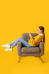 Pretty young woman using tablet computer in soft armchair on yellow background