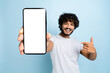 Mock-up, copy space. Happy excited indian or arabian guy, in casual wear, holds smartphone with blank white screen for presentation and points finger at it, stands on isolated blue background, smile