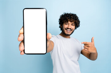Mock-up, copy space. Happy excited indian or arabian guy, in casual wear, holds smartphone with blank white screen for presentation and points finger at it, stands on isolated blue background, smile