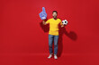 © ViDi Studio - Full size body length young bearded man football fan in yellow t-shirt cheer up support favorite team hold soccer ball fan foam glove finger up isolated on plain dark red background studio portrait.