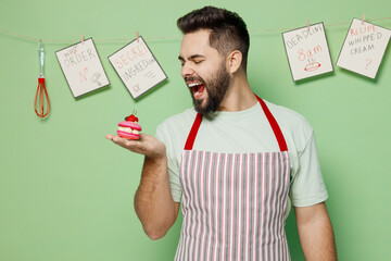 Canvas Print - Young happy male chef confectioner baker man 20s in striped apron holding bite pink little cake muffin macaroon isolated on plain pastel light green background studio portrait. Cooking food concept.