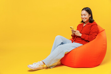 Full size body length vivid charming fun young woman of Asian ethnicity 20s in casual clothes sit in bag chair hold in hand use mobile cell phone isolated on plain yellow background studio portrait.