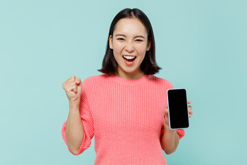 Wall Mural - Young happy woman of Asian ethnicity 20s wear pink sweater hold in hand use mobile cell phone with blank screen workspace area do winner gesture isolated on pastel plain light blue background studio.