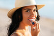 © Syda Productions - people, summer and swimwear concept - portrait of happy smiling young woman in bikini swimsuit and straw hat applying lip balm on beach