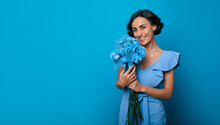 Young Gorgeous Woman In A Bright Blue Dress Is Looking In The Camera With A Big Smile, Holding A Bunch Of Blue Flowers In Her Hands. International Women's Day