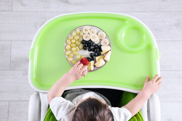 Poster - Cute little baby eating healthy food in high chair indoors, top view