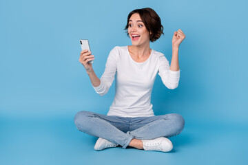 Photo of lucky shiny lady dressed white shirt legs crossed chatting modern device rising fist isolated blue color background