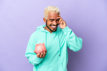 Canvas Print - Young Colombian man holding a piggybank isolated on purple background laughing