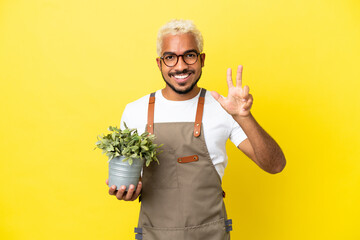 Wall Mural - Young Colombian man holding a plant isolated on yellow background happy and counting three with fingers