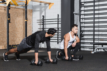 Wall Mural - Sportsman holding dumbbell near arabian friend in gym.