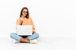 © luismolinero - Young woman with a laptop sitting on the floor smiling and showing victory sign