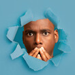 © Prostock-studio - Surprised young african american guy with wide open eyes closed his mouth with hands, looks through hole in blue paper