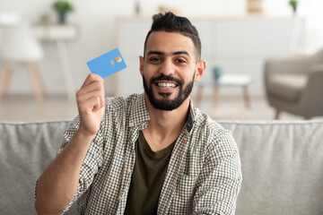 Wall Mural - Portrait of positive young Arab man showing credit card, shopping from home