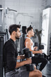 © LIGHTFIELD STUDIOS - Side view of arabian sportsman holding dumbbells near blurred friend in sports center.