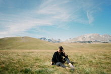 Man Sits On A Lawn Against The Backdrop Of Mountains, Turning His Head To The Side