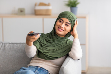 Canvas Print - Young Arab woman in Muslim hijab holding remote control, watching TV on sofa at home