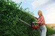 © serhiibobyk - Pretty young woman with blond hair trimming bushes outdoors. Female gardener gloves using hand electric machine. Landscaping process.