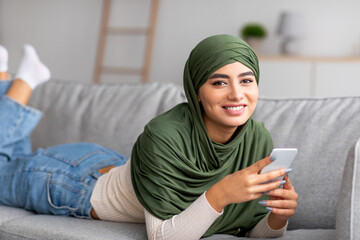 Wall Mural - Arab woman in hijab lying on sofa, using mobile phone, checking social network, browsing web, communicating online