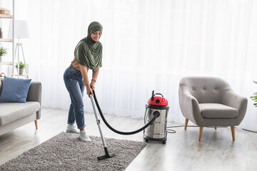 Wall Mural - Cheerful muslim woman cleaning rug carpet with vacuum cleaner