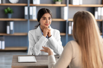 Wall Mural - Attentive cheerful pretty cute young indian female therapist in white coat listen client in clinic office