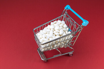 Wall Mural - Shopping cart filled with white pills and tablets on red background. Purchasing medicine. Health care business concept.