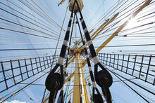 A View From Below Of The Huge Mast Of An Old Sailing Ship, Many Ropes Hanging Down From Above, Blue Sky In The Background On A Sunny Day