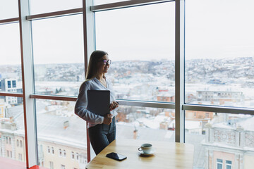 Cheerful young fair-haired business woman of European appearance in a white shirt and glasses holds a portable laptop in her hands, stands near a large window in the office on the top floor