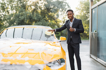 Canvas Print - Handsome young African American businessman cleaning his car windscreen with yellow sponge and soap foam outdoors at car wash service and showing thumb up