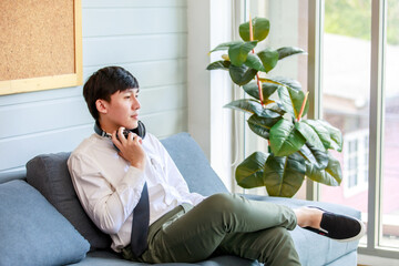 Asian young handsome professional successful male businessman employee in formal business shirt and necktie sitting on cozy sofa listening to streaming music online browsing internet with smartphone