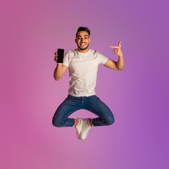 Poster - Funky young Arab guy jumping and showing modern smartphone with empty screen in neon light, mockup